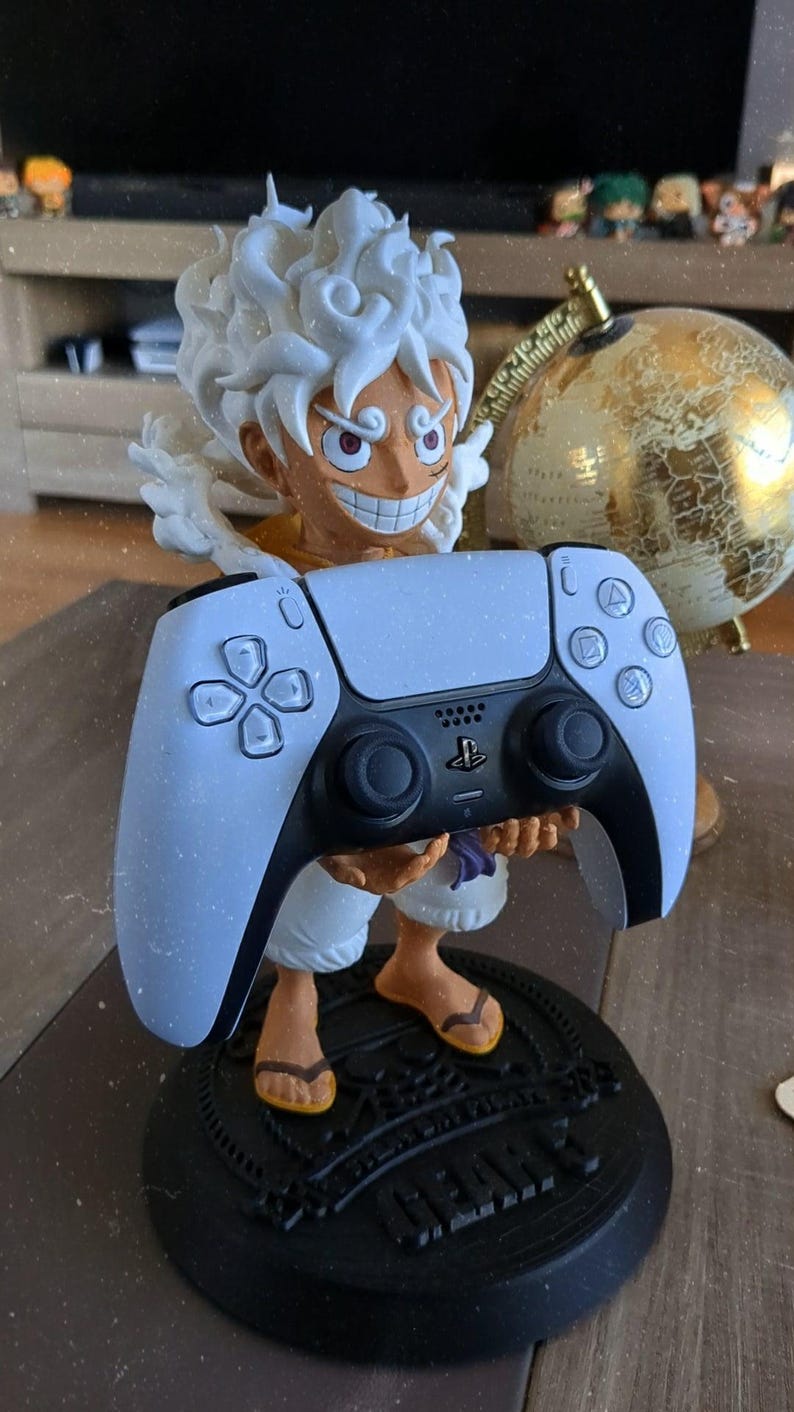 May include: A figurine of a character with spiky white hair and a wide grin, standing on a black circular base with the text GEAR 5. The character is holding a white and black PlayStation 5 game controller in their hands, positioned in front.