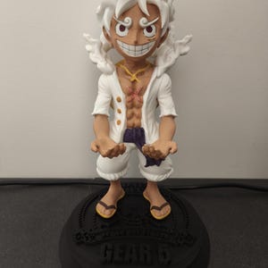 May include: A figurine of a character with spiky white hair, a wide grin, and large eyes. The figure wears an open white shirt, white shorts, and a purple sash, standing on a black circular base with the text "GEAR 5" and "STRAW HAT PIRATE" printed on it.
