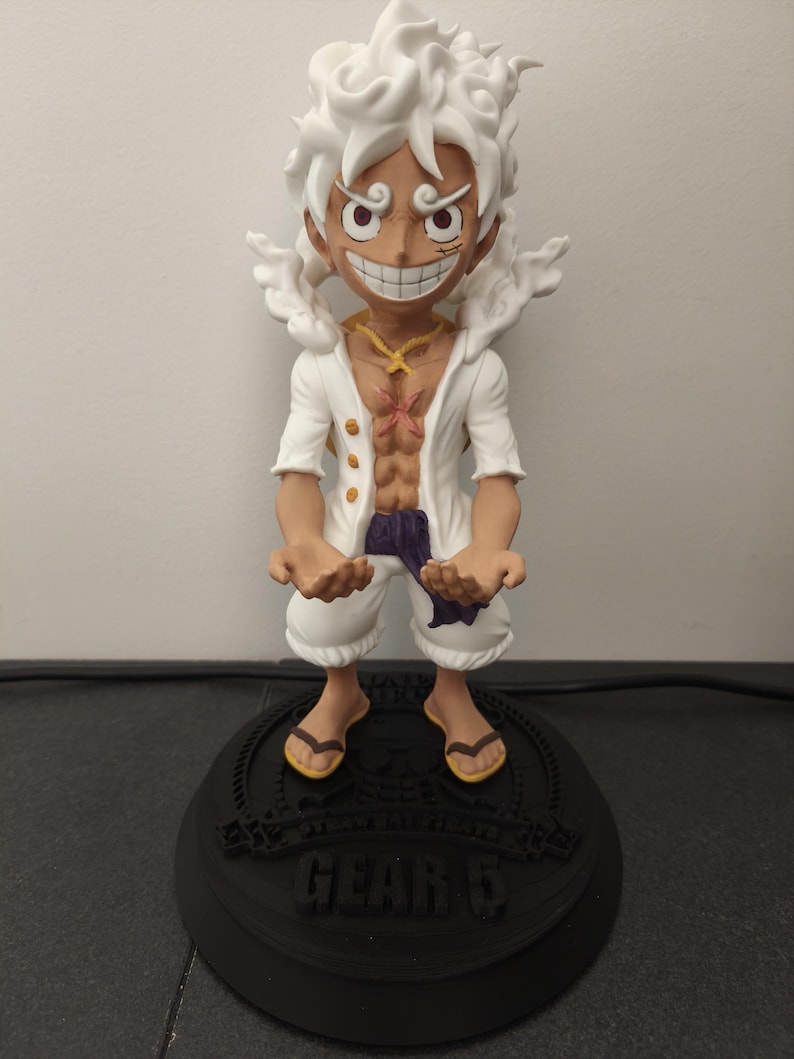 May include: A figurine of a character with spiky white hair, a wide grin, and large eyes. The figure wears an open white shirt, white shorts, and a purple sash, standing on a black circular base with the text "GEAR 5" and "STRAW HAT PIRATE" printed on it.