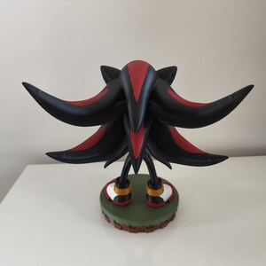 Peut inclure: Figurine noire et rouge de Shadow the Hedgehog, personnage de la s&eacute;rie de jeux vid&eacute;o Sonic the Hedgehog. La figurine pr&eacute;sente un design d&eacute;taill&eacute;, avec des pointes noires et des accents rouges. Elle repose sur une base verte avec une bordure marron.