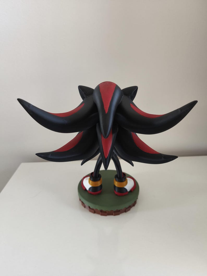 Peut inclure: Figurine noire et rouge de Shadow the Hedgehog, personnage de la s&eacute;rie de jeux vid&eacute;o Sonic the Hedgehog. La figurine pr&eacute;sente un design d&eacute;taill&eacute;, avec des pointes noires et des accents rouges. Elle repose sur une base verte avec une bordure marron.