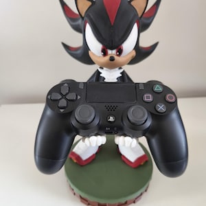 Peut inclure: Figurine noire et rouge de Shadow the Hedgehog tenant une manette noire PlayStation 4. La figurine est sur une base verte avec une bordure marron. La manette a une disposition de boutons standard.