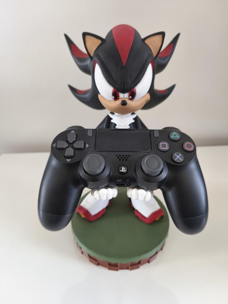 Peut inclure: Figurine noire et rouge de Shadow the Hedgehog tenant une manette noire PlayStation 4. La figurine est sur une base verte avec une bordure marron. La manette a une disposition de boutons standard.
