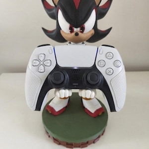 Peut inclure: Une figurine noire, rouge et blanche de Shadow the Hedgehog tenant une manette blanche de PlayStation 5. La figurine est pos&eacute;e sur une base verte avec un bord marron. La manette a des boutons noirs et des poign&eacute;es blanches textur&eacute;es.