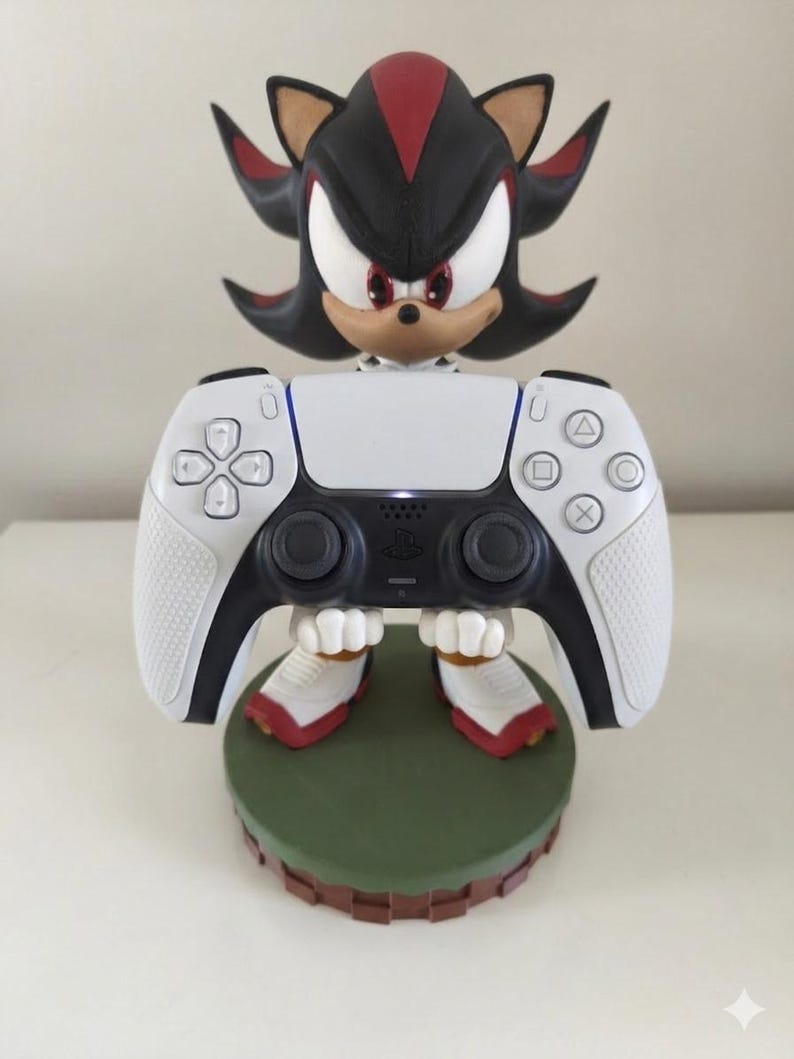 Peut inclure: Une figurine noire, rouge et blanche de Shadow the Hedgehog tenant une manette blanche de PlayStation 5. La figurine est pos&eacute;e sur une base verte avec un bord marron. La manette a des boutons noirs et des poign&eacute;es blanches textur&eacute;es.