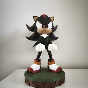 Peut inclure: Figurine de Shadow the Hedgehog, un h&eacute;risson noir avec des accents rouges, des gants blancs et des chaussures rouges et blanches. La figurine est pos&eacute;e sur une base verte et marron, avec un fond neutre.