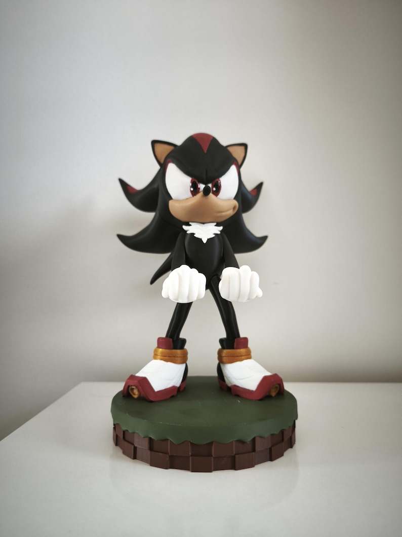 Peut inclure: Figurine de Shadow the Hedgehog, un h&eacute;risson noir avec des accents rouges, des gants blancs et des chaussures rouges et blanches. La figurine est pos&eacute;e sur une base verte et marron, avec un fond neutre.