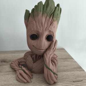 Peut inclure: Une sculpture détaillée de Groot, un personnage des Gardiens de la Galaxie. La figurine est principalement marron avec des accents verts sur la tête. Groot est posé avec sa main sur son visage, souriant. La sculpture est sur une surface en bois.