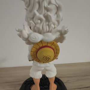 May include: A figurine of a character with spiky white hair and a cloud-like aura, wearing white shorts and a yellow straw hat with a red band on the back. The figure stands on a circular black base with embossed details, viewed from the rear.