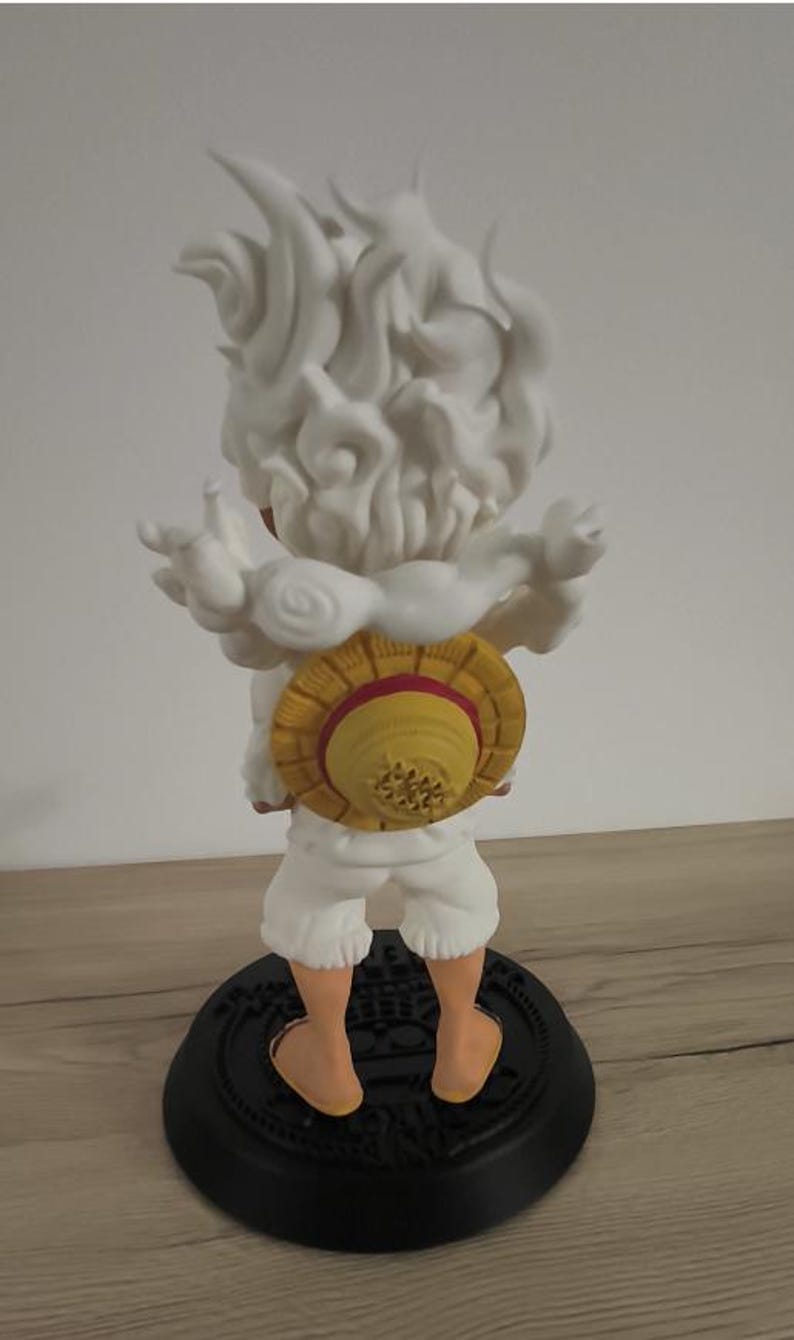 May include: A figurine of a character with spiky white hair and a cloud-like aura, wearing white shorts and a yellow straw hat with a red band on the back. The figure stands on a circular black base with embossed details, viewed from the rear.