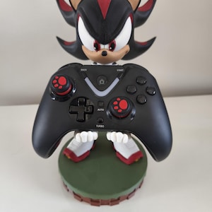 Peut inclure: Une manette de jeu noire tenue par une figurine de Shadow the Hedgehog. La manette a des accents rouges sur les joysticks et les boutons. La figurine est noire, rouge et blanche, et repose sur une base verte.