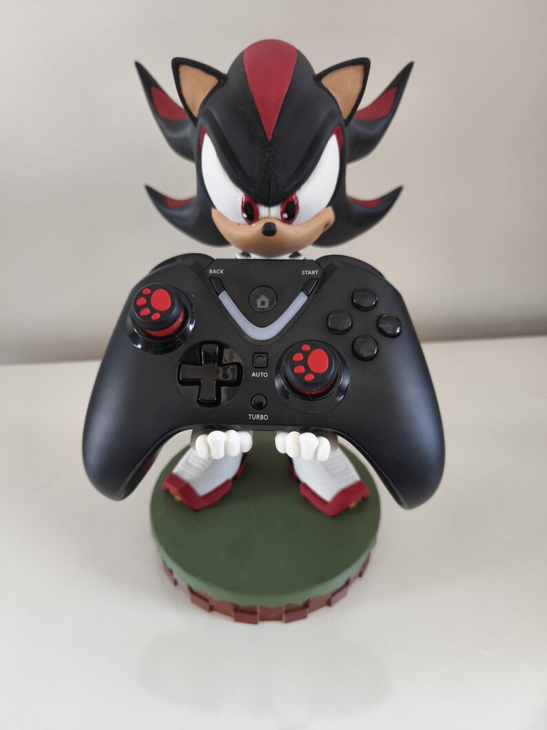 Peut inclure: Une manette de jeu noire tenue par une figurine de Shadow the Hedgehog. La manette a des accents rouges sur les joysticks et les boutons. La figurine est noire, rouge et blanche, et repose sur une base verte.