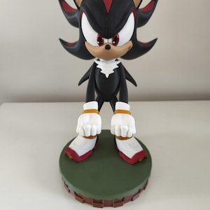 Peut inclure: Figurine d&eacute;taill&eacute;e de Shadow the Hedgehog, personnage de la franchise Sonic the Hedgehog. La figurine est principalement noire avec des accents rouges et blancs, pos&eacute;e sur une base verte et marron. Le personnage a des yeux blancs et une expression d&eacute;termin&eacute;e.