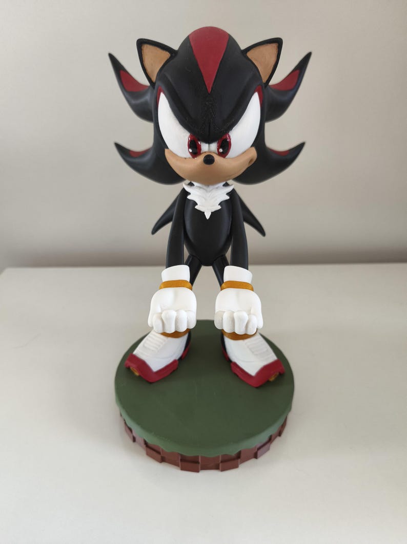 Peut inclure: Figurine d&eacute;taill&eacute;e de Shadow the Hedgehog, personnage de la franchise Sonic the Hedgehog. La figurine est principalement noire avec des accents rouges et blancs, pos&eacute;e sur une base verte et marron. Le personnage a des yeux blancs et une expression d&eacute;termin&eacute;e.
