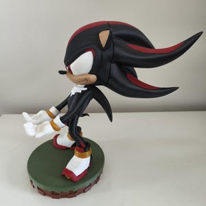 Peut inclure: Figurine noire et rouge d'un personnage avec des gants et des chaussures blancs, debout sur une base verte. Le personnage a un visage blanc, un museau beige et des &eacute;pines rouges et noires. La figurine est pos&eacute;e avec les bras tendus.