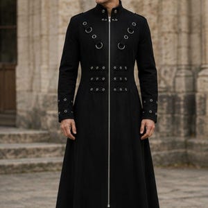 May include: A long black coat featuring a high collar, a full-length silver front zip, and decorative silver metal rings and studs arranged in rows across the chest and cuffs. The garment has long sleeves and a structured, floor-length silhouette.