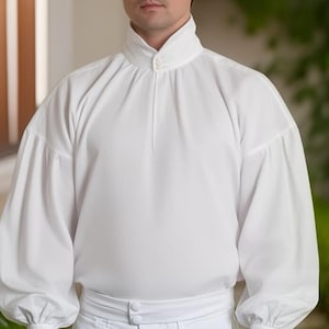 Può includere: Una camicia bianca a maniche lunghe con colletto alto a due bottoni, maniche voluminose arricciate e un girovita aderente. Il capo è realizzato in un tessuto bianco liscio e uniforme, caratterizzato da un design pulito e strutturato.