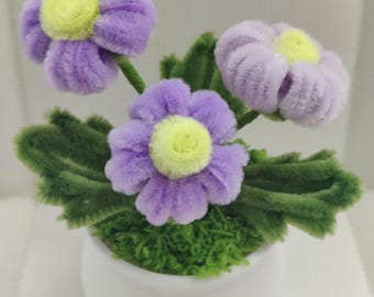 May include: A whimsical floral arrangement featuring three purple pipe cleaner flowers with yellow centers. The flowers sit atop green leaves and moss in a white pot with a smiling face design. A charming, handmade decorative item.