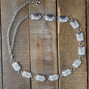 May include: A silver-toned chain belt featuring a series of repeating rectangular metal plates with rounded corners and engraved geometric patterns. The belt includes a long adjustable chain section with a circular ring at the end for closure.
