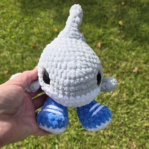 May include: A handmade, gray crocheted shark plushie with black eyes and small gray arms. The shark is wearing blue and white striped sneakers. The plushie is held in front of a green grassy background.