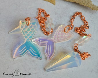 Puede incluir: Dos péndulos con forma de cola de sirena iridiscente con una cadena de cobre y un péndulo de punta de cristal transparente iridiscente. Los péndulos están sobre un fondo blanco.