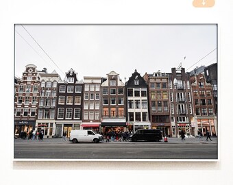 May include: A framed photograph of a street scene in Amsterdam, featuring a row of historic buildings with unique facades. The image captures the architecture, with people and vehicles on the street below a cloudy sky.
