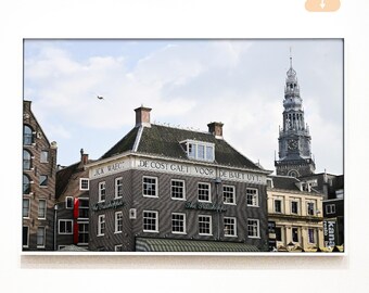 May include: A framed photograph of a historic building in Amsterdam, Netherlands. The building is gray brick with white-framed windows and text above the windows. A tall clock tower is visible in the background under a cloudy sky.