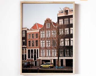 May include: A framed print featuring a row of historic buildings in Amsterdam. The buildings are various shades of brown and black, with red tile roofs. A yellow car is parked on the street in front of the buildings.