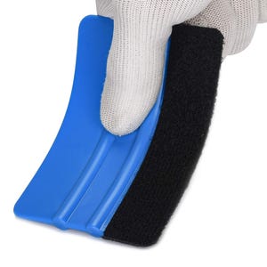 Puede incluir: Una espátula de plástico azul con un borde de fieltro negro es sostenida por una mano enguantada. La espátula tiene una forma curva con tres ranuras paralelas. El fieltro negro es rectangular y está adherido a la espátula.