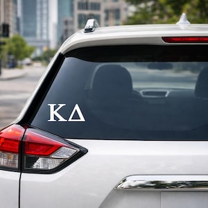 May include: White car window decal featuring the Greek letters ΚΔ. The decal is in a sans-serif font and is centered on the rear window of a white SUV. The background shows a city street with buildings and trees.
