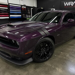Puede incluir: Un Dodge Challenger color ciruela con franjas negras de carreras y ruedas negras. El coche est&aacute; estacionado en un garaje con rollos de vinilo en la pared. El coche tiene una toma de aire en el cap&oacute; y un aler&oacute;n delantero.