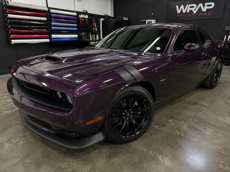 Puede incluir: Un Dodge Challenger color ciruela con franjas negras de carreras y ruedas negras. El coche est&aacute; estacionado en un garaje con rollos de vinilo en la pared. El coche tiene una toma de aire en el cap&oacute; y un aler&oacute;n delantero.