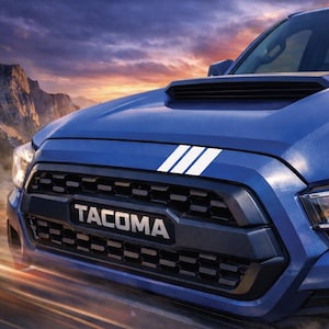 May include: Close-up of a blue Tacoma truck. The front grill is black with the name "Tacoma" in silver lettering. The hood has three white stripes. The background shows a mountain range and a sunset.