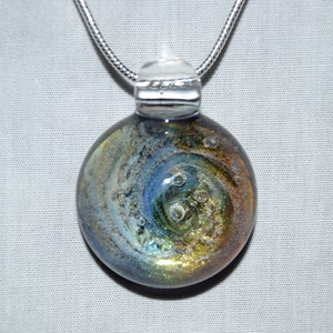 May include: A silver chain necklace with a round glass pendant. The pendant features a swirling pattern of blue, gold, and white with small air bubbles trapped inside.