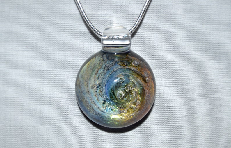 May include: A silver chain necklace with a round glass pendant. The pendant features a swirling pattern of blue, gold, and white with small air bubbles trapped inside.
