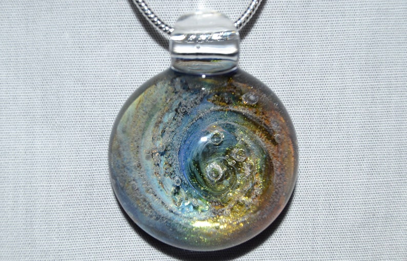 May include: A round glass pendant with a silver chain. The glass is a swirling mix of blue, green, and gold with small air bubbles trapped inside.