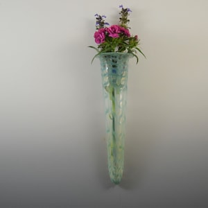 May include: A light blue glass vase with a white and green pattern. The vase is tall and narrow, and it is filled with pink and purple flowers.