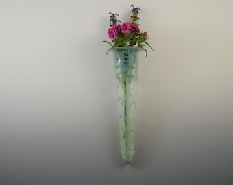 Puede incluir: Un jarrón de vidrio azul claro con un patrón blanco y verde. El jarrón es alto y estrecho, y está lleno de flores rosas y moradas.