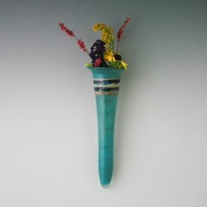May include: A turquoise glass vase with a white and blue stripe design, holding a bouquet of yellow, red, and purple flowers.