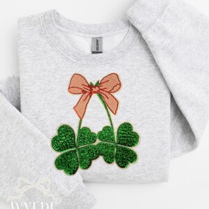 May include: A gray sweatshirt with a green and pink sequin patch of a four-leaf clover with a bow on top. The sweatshirt has the text "WYLDE & OZZY" on the bottom left corner.