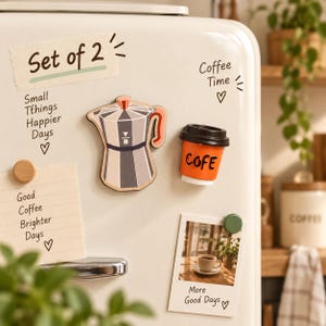 May include: A set of two refrigerator magnets featuring a gray and white moka pot with a coral handle and an orange coffee cup with a black lid and the text COFE. The magnets are displayed on a cream-colored refrigerator surface with handwritten notes.