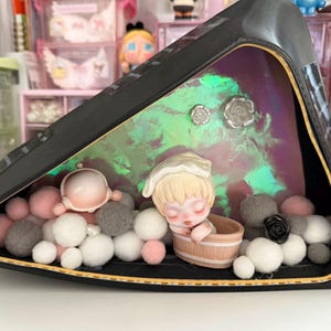 May include: A triangular black display case with a sleeping doll inside. The doll is wearing a white hat and has blonde hair. The case is filled with white, pink, and gray pom-poms. The background is a iridescent green and purple.