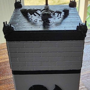 May include: A black and grey 3D printed dice tower with a brick pattern and a cross on top. The tower has a skull and a teardrop symbol on the front.