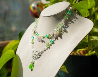 May include: A two-strand necklace with a nature-inspired design. The necklace features silver chains with green beads, white pearls, and small floral accents. A silver leaf pendant hangs below, with a green crystal drop. The necklace is displayed on a white bust.
