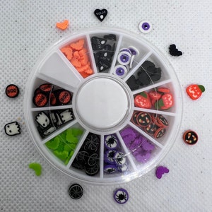 May include: A white plastic wheel with 12 compartments filled with various Halloween-themed nail art charms. The charms include ghosts, bats, pumpkins, spiders, candy corn, and eyeballs. The wheel is on a white surface.