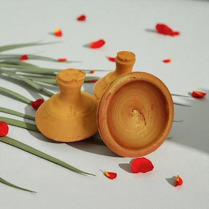 Puede incluir: Tres pequeñas ollas de arcilla hechas a mano, de color terracota. Dos tienen forma de botella con tapones tipo corcho, y una es un cuenco poco profundo. Pétalos de rosa rojos y hojas verdes están esparcidos alrededor de las ollas sobre una superficie blanca.