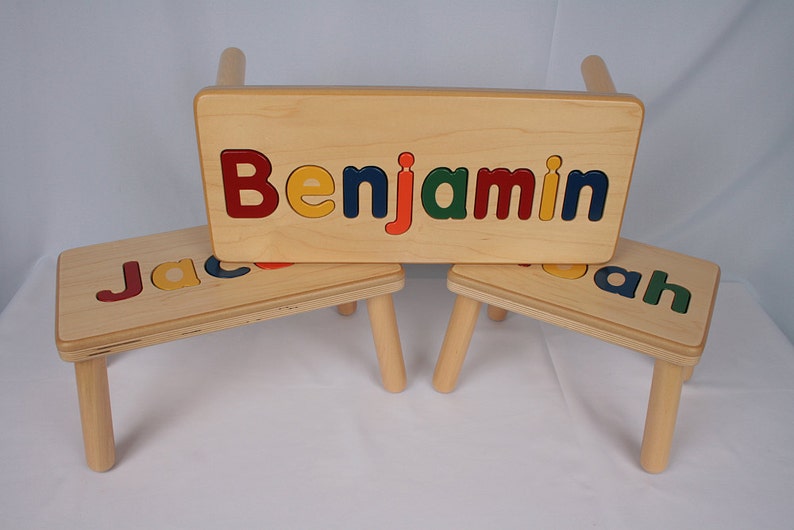 May include: Three wooden children's stools with personalized names spelled out in colorful letters. The stools are made of natural wood and have a simple design. The names on the stools are "Benjamin", "Jacob", and "Noah".