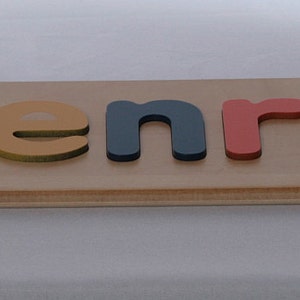 May include: Wooden puzzle with the name "Henry" spelled out in colorful letters. The letters are red, blue, yellow, and green. The letters are placed in the slots of the wooden board.