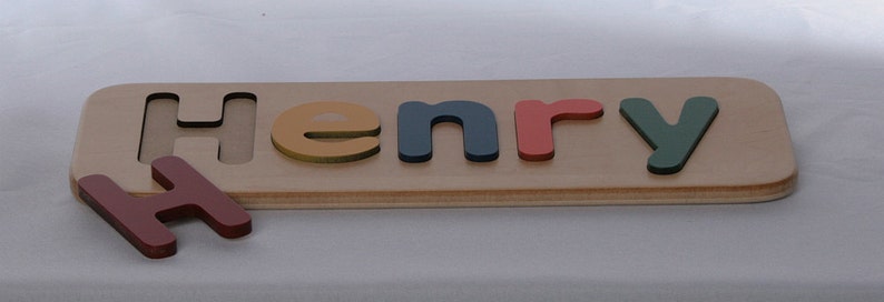 May include: Wooden puzzle with the name "Henry" spelled out in colorful letters. The letters are red, blue, yellow, and green. The letters are placed in the slots of the wooden board.