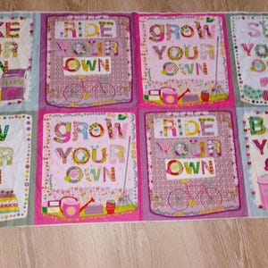 May include: A colorful fabric panel featuring eight squares with the phrases "Bake Your Own", "Ride Your Own", "Grow Your Own", and "Sew Your Own". Each square has decorative elements like cupcakes, sewing machines, and bicycles.